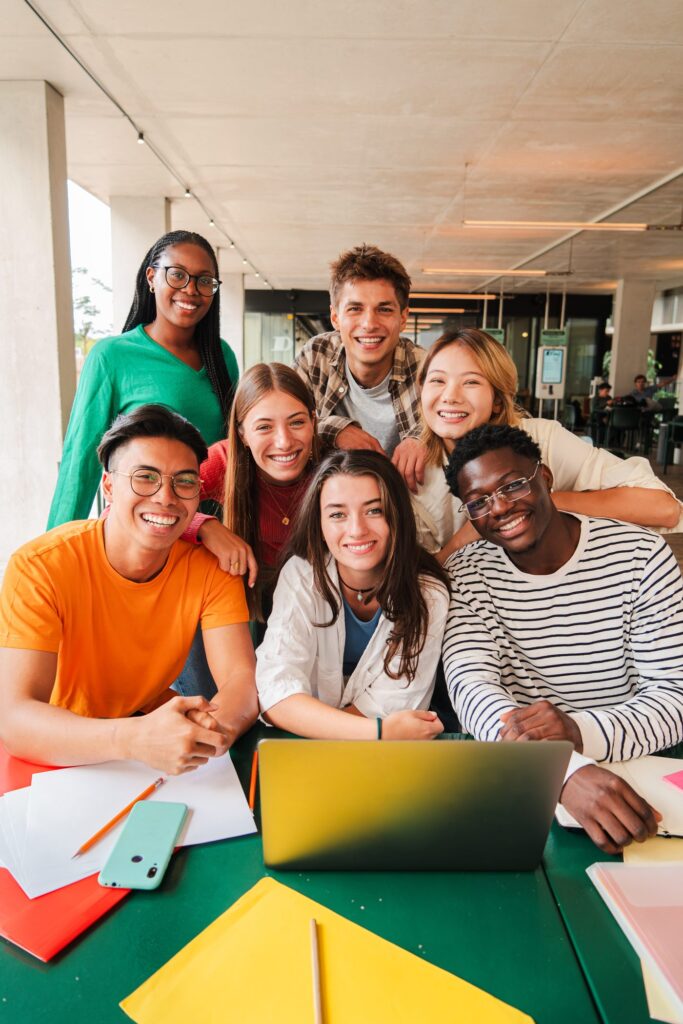A diverse group of seven young people gathered around a table, smiling together while studying with a laptop.